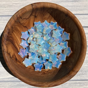 May include: A wooden bowl filled with numerous opalescent star-shaped beads. The beads have a translucent quality with hints of blue and white, creating a shimmering effect. The bowl is made of a light brown wood.