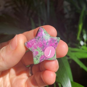 May include: A star-shaped gemstone with a pink and green mottled pattern. A small, round sticker with the letter "J" is affixed to the top of the star. The gemstone is held in a person's hand.