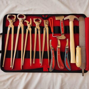 Könnte beinhalten: Eine Sammlung von Hufschmiedewerkzeugen, darunter Zangen, Hämmer, Messer und eine Raspel, angeordnet in einer rot-schwarzen Werkzeugrolle. Die Werkzeuge haben Holz- und Metallgriffe.