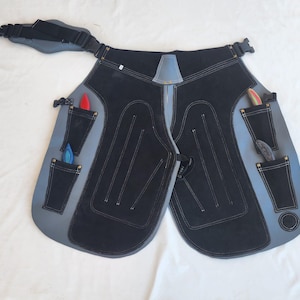May include: Black and gray leather farrier apron with multiple pockets and a waist strap. The apron has a black suede-like center panel, gray edges, and white stitching. Several tools are visible in the pockets.