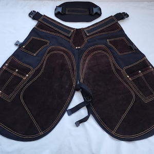 Könnte beinhalten: Schwarze und braune Hufschmiedschürze mit Kniepolstern und Taschen aus Wildleder. Die Schürze hat orangefarbene Nähte und verstellbare Riemen. Eine schwarze Taillenstütze ist enthalten.