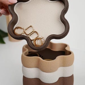 May include: A stackable, flower-shaped jewellery organiser with a dark brown lid featuring a cream-coloured mesh insert. The lid is held up to reveal gold hoop and square earrings inside. The base consists of stacked beige, white, and brown segments.