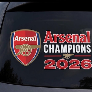 May include: A car window decal featuring the Arsenal team logo and the words "Arsenal Champions 2026." The logo is a red shield with a gold cannon and the team name. The text is in red and white.