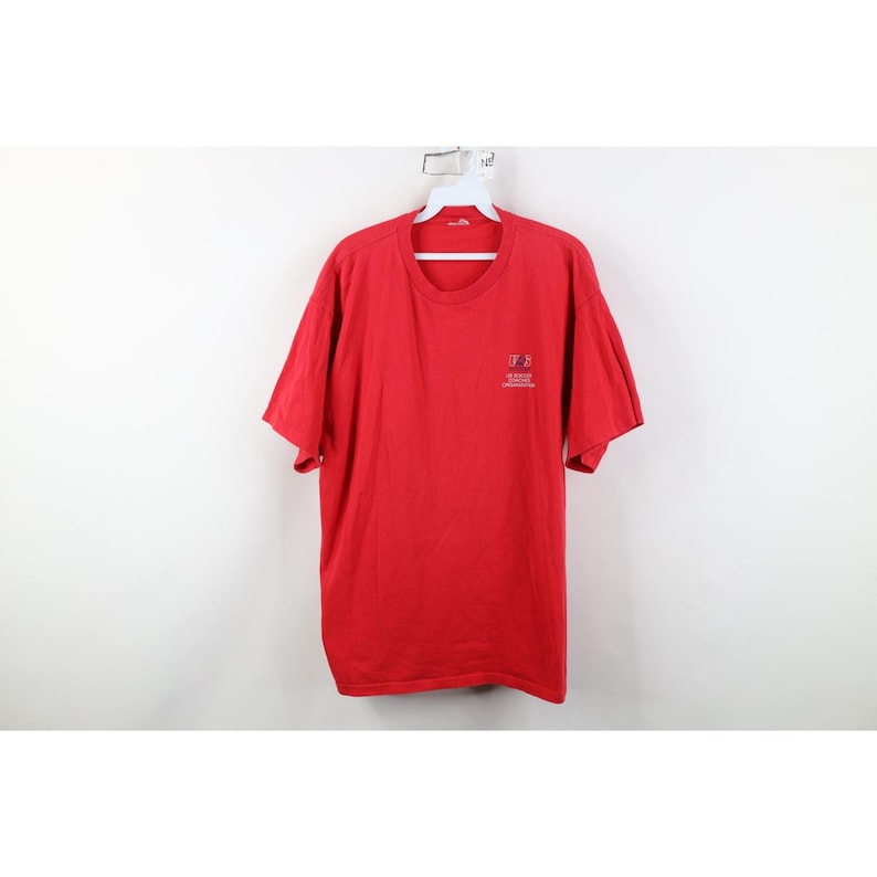 May include: A red short-sleeve t-shirt with a small logo on the upper left chest. The shirt is hanging on a white hanger against a white background. The t-shirt is made of a soft material.