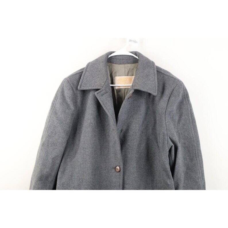 May include: A gray wool coat with a classic collar and button closure. The coat is hanging on a white hanger against a white background. The coat has a tailored fit and a simple, elegant design.
