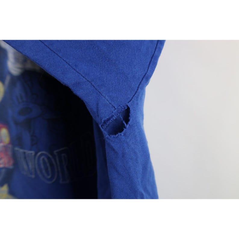 May include: Close-up of a royal blue t-shirt with a distressed design. The shirt has a torn section near the shoulder. The word "WOR" is visible on the shirt. The shirt appears to be vintage.