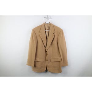 May include: A solid tan blazer featuring a notched lapel, two front buttons, and two flap pockets at the waist. The garment has long sleeves and is displayed on a white plastic hanger against a plain, neutral background for clear visibility.