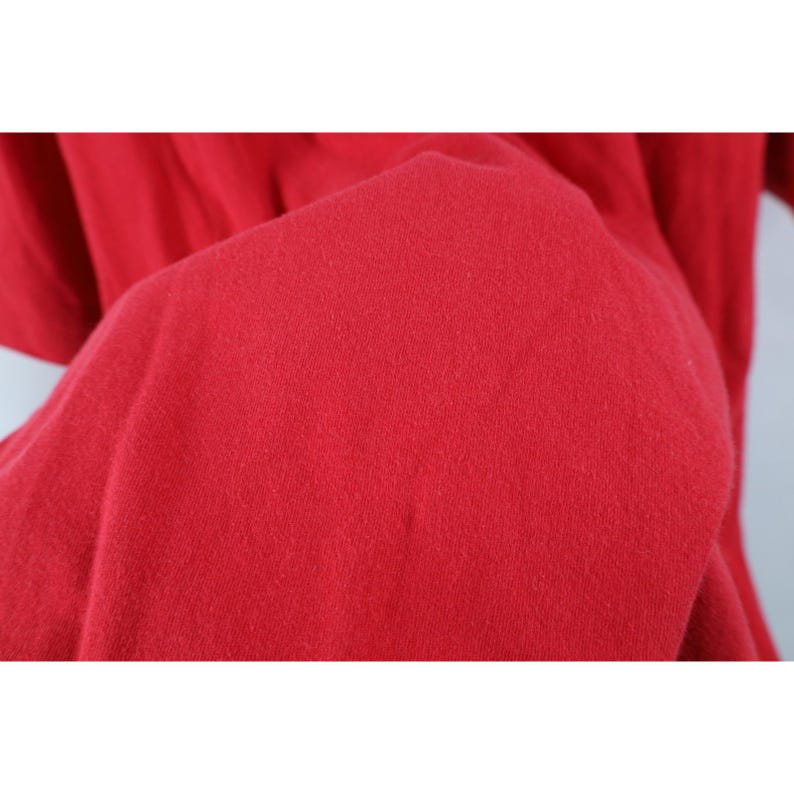 May include: Close-up of a vibrant red fabric, likely a piece of clothing. The material appears soft and slightly textured, with subtle variations in the color. The image focuses on the texture and color of the textile.