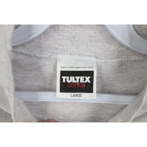 Op de afbeelding: Close-up van een grijs katoenen T-shirt met een wit label. Op het label staat "TULTEX COTTON" in witte tekst op een zwarte achtergrond, met daaronder "LARGE". Het shirt hangt aan een witte hanger.