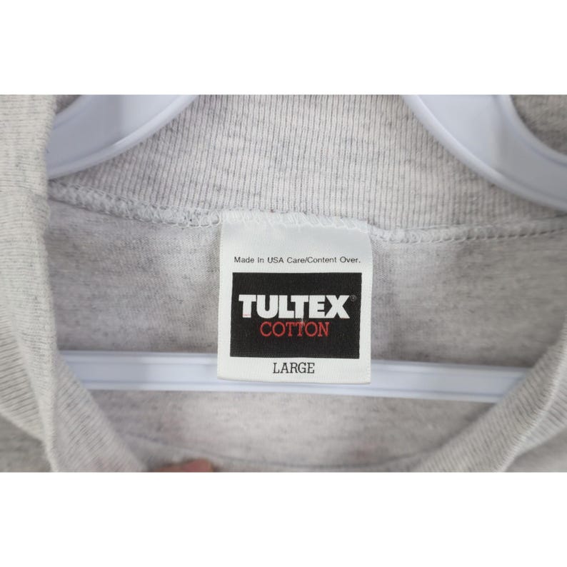 Op de afbeelding: Close-up van een grijs katoenen T-shirt met een wit label. Op het label staat "TULTEX COTTON" in witte tekst op een zwarte achtergrond, met daaronder "LARGE". Het shirt hangt aan een witte hanger.