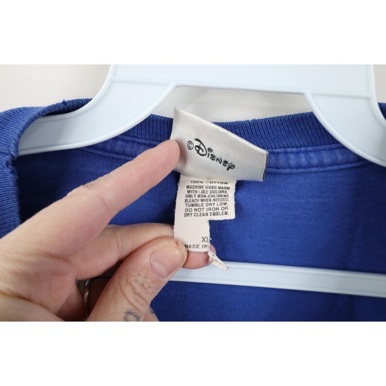 May include: A blue cotton t-shirt with a Disney label. The label includes washing instructions and the size XL. The shirt is hanging on a white hanger.