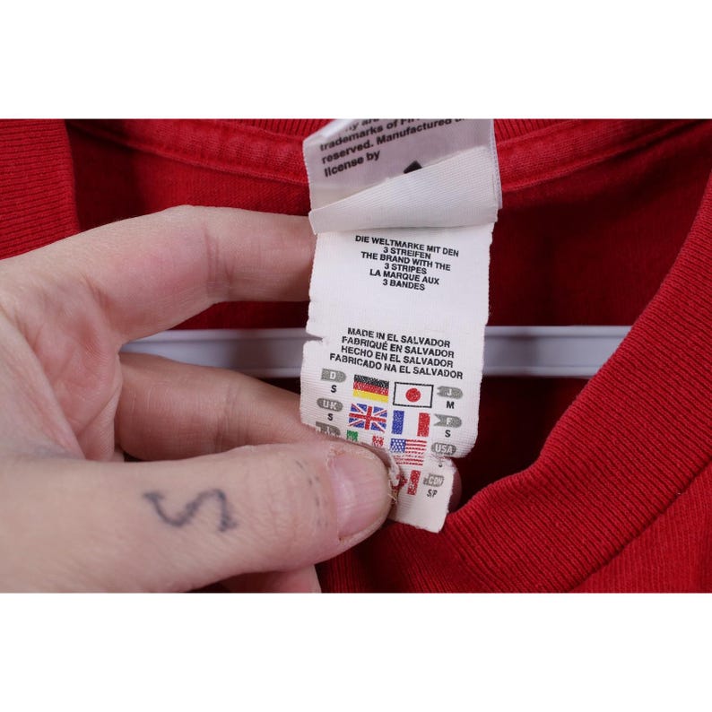 May include: Close-up of a red t-shirt with a tag. The tag includes text in multiple languages, including "DIE WELTMARKE MIT DEN 3 STREIFEN" and "THE BRAND WITH THE 3 STRIPES". Flags of different countries are also visible on the tag.