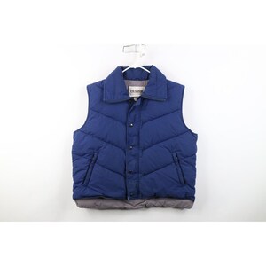 May include: A navy blue quilted vest with a collar and snap closure. The vest has two side pockets and a gray bottom hem. The brand name "EXCURSION" is visible on the collar. The vest is hanging on a white hanger.