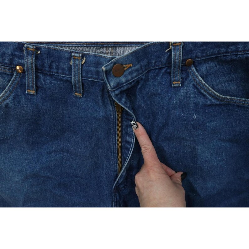 Op de afbeelding: Close-up van een blauwe spijkerbroek met een messing ritssluiting en knoopsluiting. De jeans heeft riemlussen en zakken. De ritssluiting is gedeeltelijk open en een vinger wijst naar de ritssluiting.