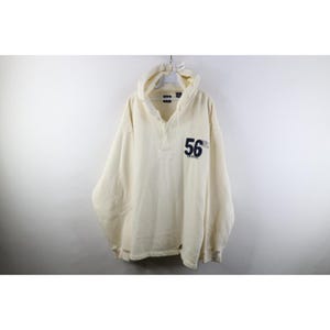 May include: A cream-coloured long-sleeve pullover hoodie featuring a partial button-up placket at the neckline. The front displays the dark blue number 56 printed on the right side. The garment includes a hood with drawstrings and ribbed cuffs.
