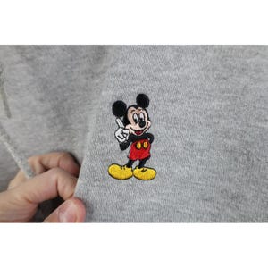 May include: A light gray sweatshirt featuring a small, detailed embroidery of Mickey Mouse. The character is depicted with black ears, a flesh-toned face, red shorts, and yellow shoes, with one white-gloved hand raised in a pointing gesture.
