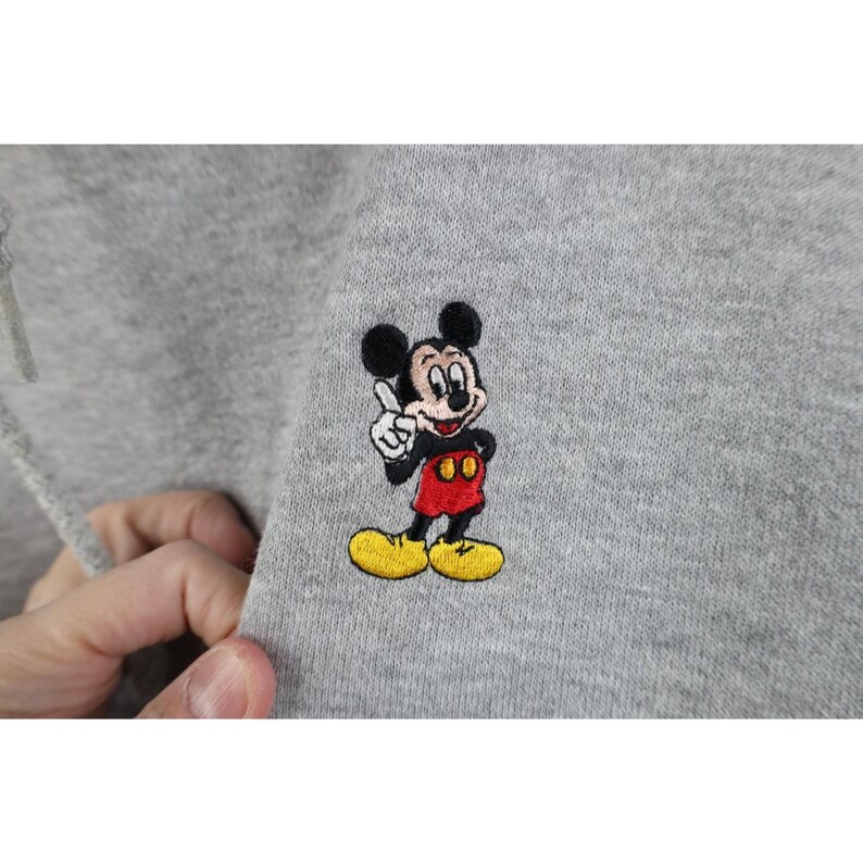 May include: A light gray sweatshirt featuring a small, detailed embroidery of Mickey Mouse. The character is depicted with black ears, a flesh-toned face, red shorts, and yellow shoes, with one white-gloved hand raised in a pointing gesture.