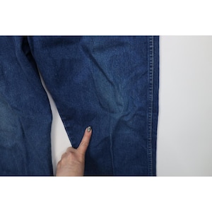 Op de afbeelding: Close-up van een blauwe spijkerbroek. De jeans heeft een medium wassing met zichtbare naden en een lichte vervaging. De stof lijkt stevig. Een hand raakt de jeans aan.