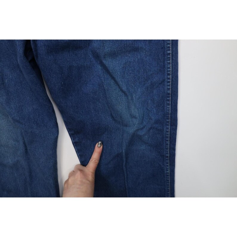 Op de afbeelding: Close-up van een blauwe spijkerbroek. De jeans heeft een medium wassing met zichtbare naden en een lichte vervaging. De stof lijkt stevig. Een hand raakt de jeans aan.