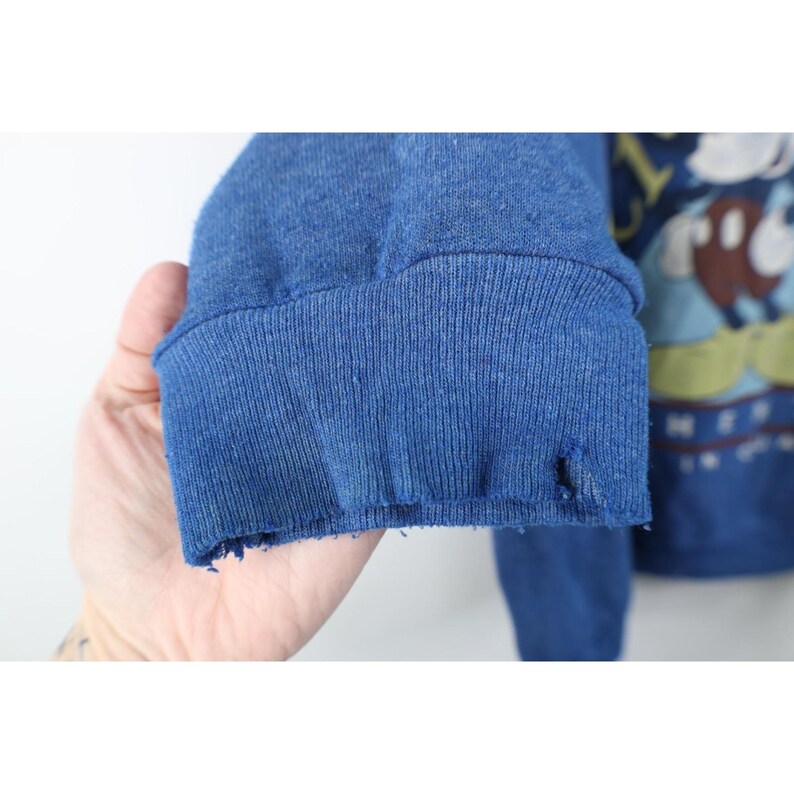 Op de afbeelding: Een blauw sweatshirt met lange mouwen en een geribbelde boord met zichtbare rafels en een klein gaatje. Het kledingstuk heeft een gedeeltelijke grafische print op de voorzijde, inclusief een figuur met bruine en witte details en gele accenten.