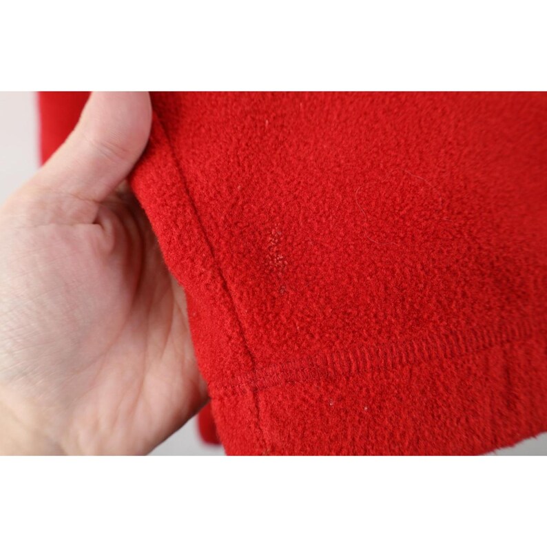 May include: Close-up of a vibrant red fleece fabric. The texture is soft and fuzzy, with visible stitching along the edge. The fabric is held by a hand, showcasing the material's warmth and texture. Ideal for clothing or blankets.