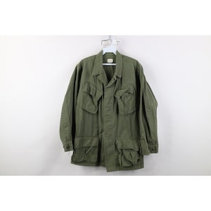 May include: An olive green military-style jacket with multiple front pockets and a collared neckline. The jacket is hanging on a hanger against a white background. The jacket has long sleeves and a button-up front.