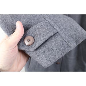 May include: Close-up of a gray wool coat sleeve with a buttoned cuff. The coat has a classic design with a large, round, brown button. The fabric appears soft and warm, suggesting a winter coat or jacket.