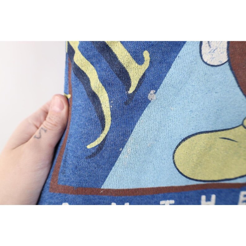 Op de afbeelding: Een blauwe sweater met een grafische print met een bruin en geel designelement in een driehoekige vorm. De stof heeft een textuur met zichtbare slijtage en een bruine rand die het onderste gedeelte van de grafische print omlijst.