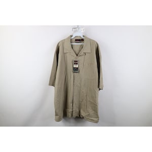 May include: A tan button-down shirt with short sleeves, hanging on a white hanger. The shirt has a collar and a pocket on the left side. A tag is attached to the shirt. The shirt is made of a light-colored fabric.