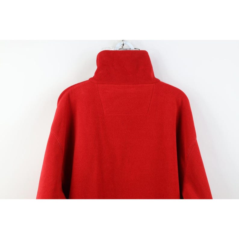 May include: A bright red fleece pullover with a high, folded collar. The garment has a simple, minimalist design with a small, rectangular patch detail on the upper back. The fleece material appears soft and warm.