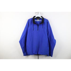 Puede incluir: Chaqueta azul de manga larga con cuello negro y cierre de cremallera de un cuarto. La chaqueta tiene un cordón en el dobladillo inferior y está colgada de una percha blanca.