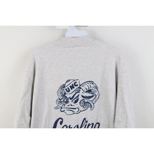 Op de afbeelding: Lichtgrijs sweatshirt met een marineblauwe afbeelding van een rammenkop met de letters "UNC" op een hoed. Het woord "Carolina" staat eronder in marineblauw. De sweatshirt heeft een ronde hals.