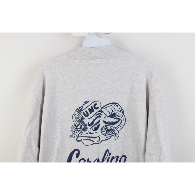 Op de afbeelding: Lichtgrijs sweatshirt met een marineblauwe afbeelding van een rammenkop met de letters "UNC" op een hoed. Het woord "Carolina" staat eronder in marineblauw. De sweatshirt heeft een ronde hals.