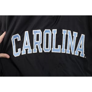 May include: Black jacket with the word "CAROLINA" in large, light blue letters outlined in white. The jacket has a small, round button at the top. The fabric appears to be a smooth, synthetic material.