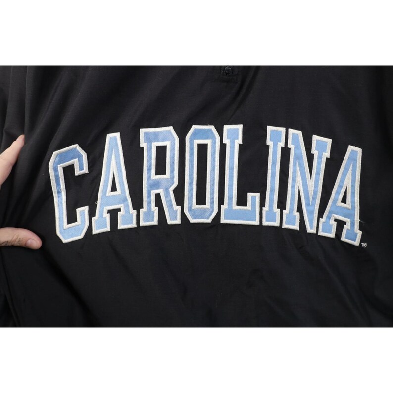 May include: Black jacket with the word "CAROLINA" in large, light blue letters outlined in white. The jacket has a small, round button at the top. The fabric appears to be a smooth, synthetic material.