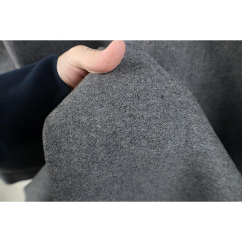 May include: Close-up of a gray fabric with a textured surface. The fabric is held by a hand, revealing a dark blue sleeve. The fabric has a few small dark spots.