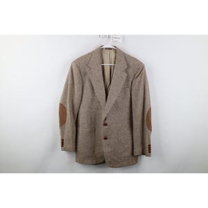 May include: A tan tweed blazer with brown elbow patches and brown buttons. The blazer is hanging on a hanger against a white background. The blazer has a classic, tailored design.