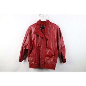 Puede incluir: Chaqueta de cuero roja con cuello alzado y cierre de cremallera frontal. La chaqueta tiene un corte holgado con detalles fruncidos en la cintura y los puños. Cuelga de una percha blanca sobre un fondo blanco.