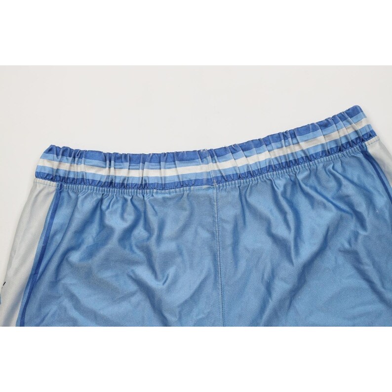 May include: Light blue athletic shorts featuring a wide, elasticized waistband with horizontal stripes in shades of white and varying tones of blue. The fabric has a smooth, slightly reflective finish, and the sides include vertical white panels.