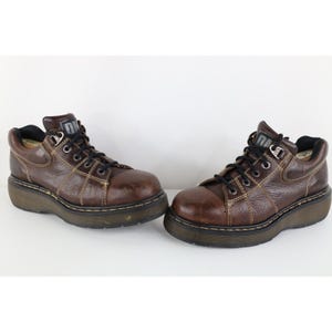 Può includere: Un paio di stivaletti in pelle marrone con lacci neri e una suola spessa e scura. Gli stivali hanno cuciture a vista e un logo sulla linguetta. Le scarpe sono su sfondo bianco.