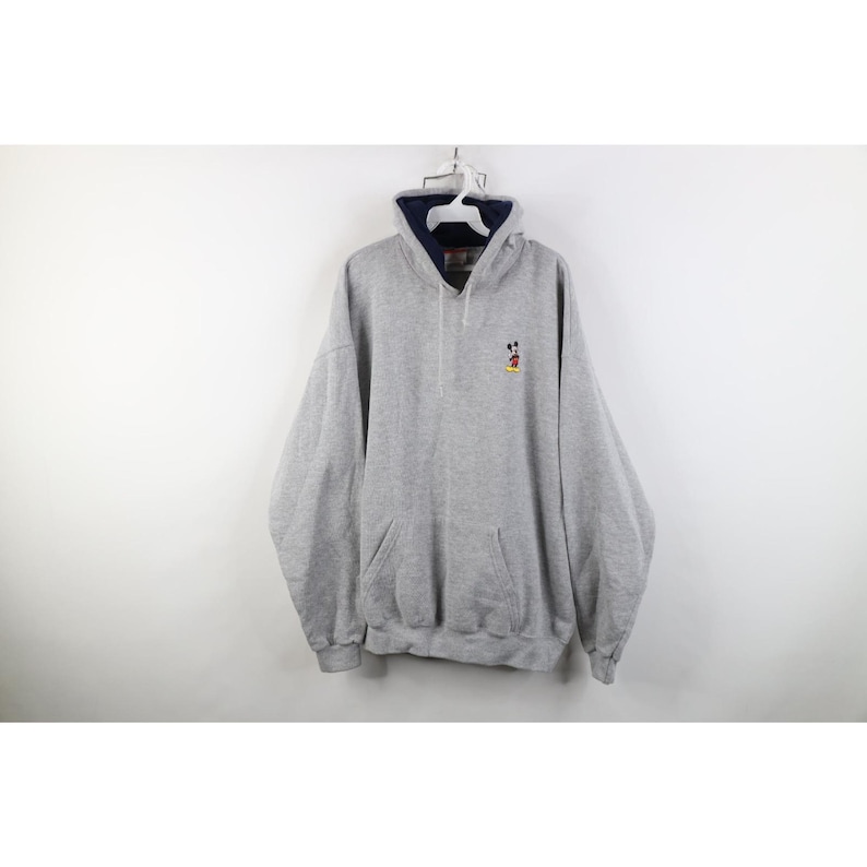 May include: A heather gray pullover hoodie featuring a small embroidered Mickey Mouse character on the left chest. The garment includes a dark navy blue inner hood lining, a front kangaroo pocket, long sleeves, and a drawstring hood closure.