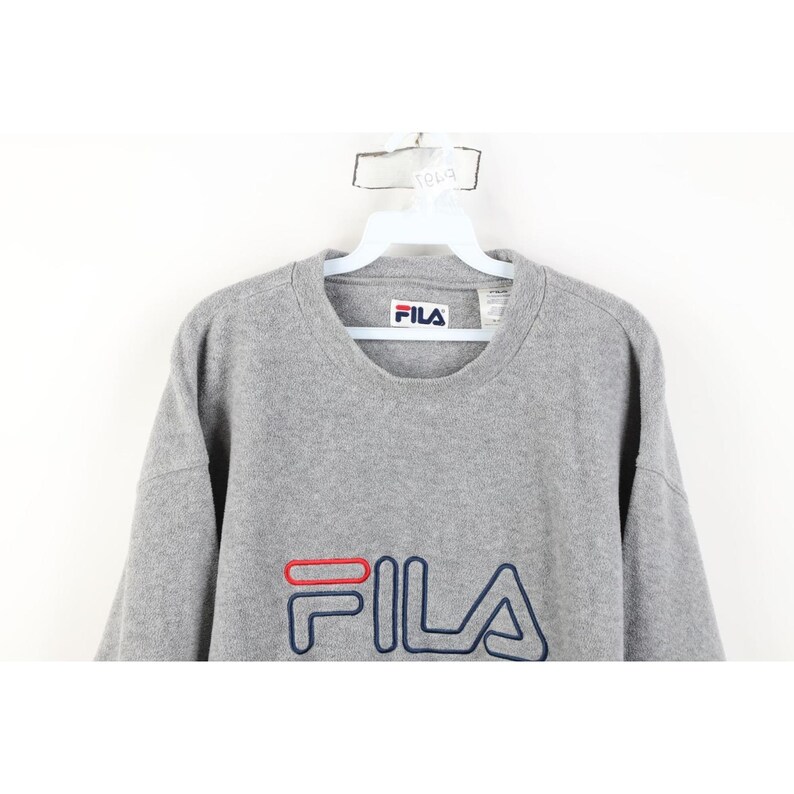 May include: A gray crew neck sweatshirt with the brand name "FILA" embroidered in red and navy blue. The sweatshirt is hanging on a white hanger against a white background.