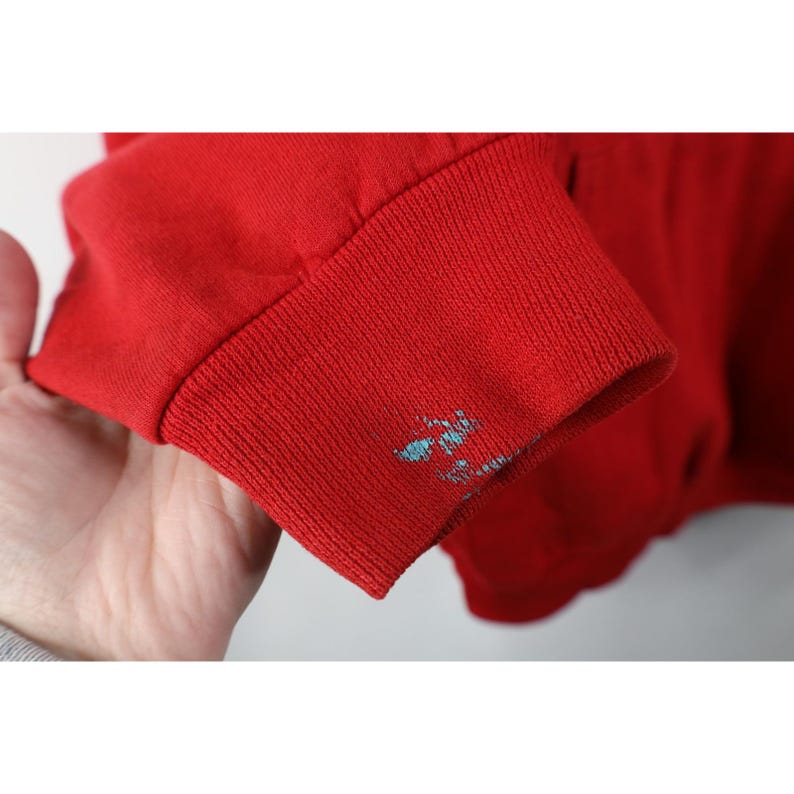Peut inclure: Gros plan sur une manche de sweat-shirt rouge avec un poignet c&ocirc;tel&eacute;. Le poignet pr&eacute;sente une petite zone d'&eacute;claboussures de peinture bleu clair. Le tissu est de couleur rouge vif.