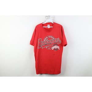May include: A red t-shirt with the text "Ohio State Buckeyes" in a stylized design. The design includes the word "Buckeyes" in a bold, arched font, with the Ohio State logo below. The shirt is hanging on a white hanger.