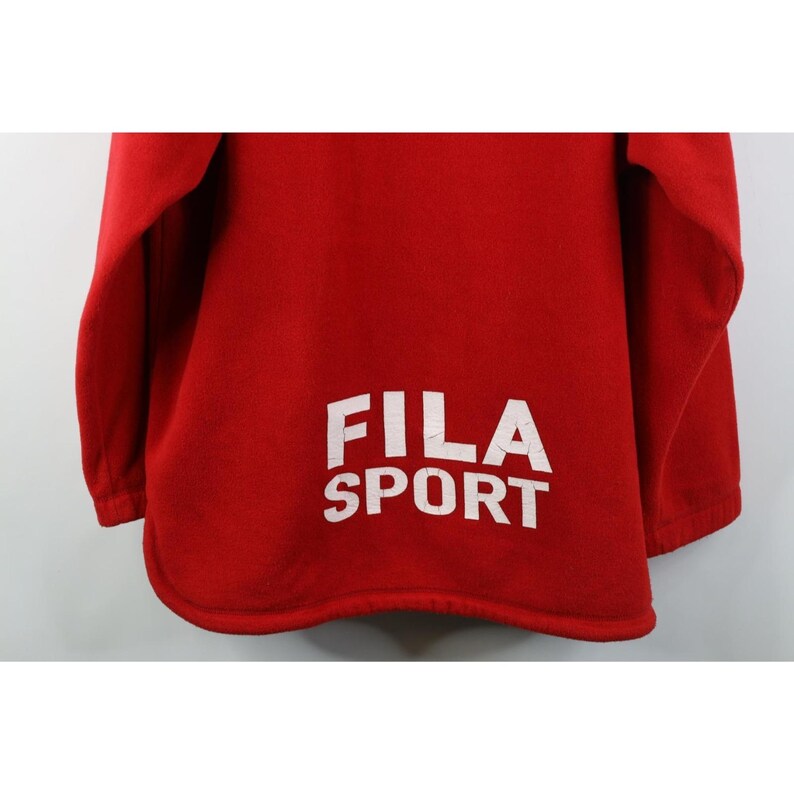 May include: A red fleece sweatshirt with the white text "FILA SPORT" printed on the lower back. The sweatshirt has long sleeves and a rounded hemline. The fabric appears soft and warm.
