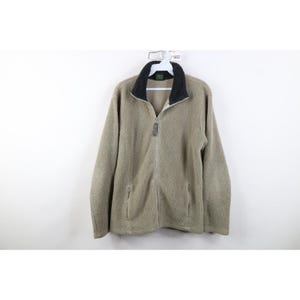 Puede incluir: Chaqueta de forro polar color beige con cuello negro y cremallera completa. La chaqueta tiene dos bolsillos laterales y un aspecto suave y texturizado. La chaqueta está colgada de una percha blanca.