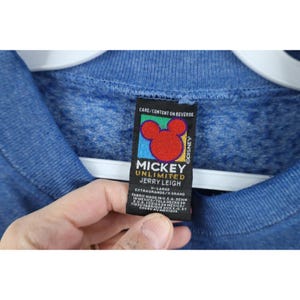 Op de afbeelding: Een blauw gem&ecirc;leerd sweatshirt met een zwart label aan de binnenzijde. Het label bevat een rood Mickey Mouse-hoofdpictogram, de tekst Mickey Unlimited Jerry Leigh, maat X-Large en informatie over de stof en de productielocatie.