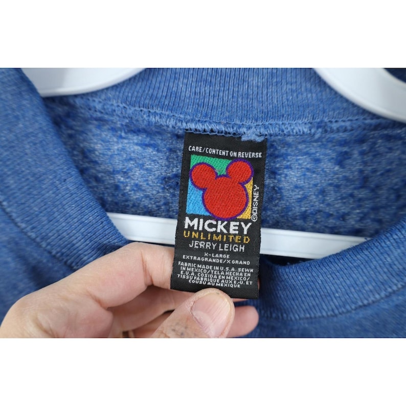 Op de afbeelding: Een blauw gem&ecirc;leerd sweatshirt met een zwart label aan de binnenzijde. Het label bevat een rood Mickey Mouse-hoofdpictogram, de tekst Mickey Unlimited Jerry Leigh, maat X-Large en informatie over de stof en de productielocatie.