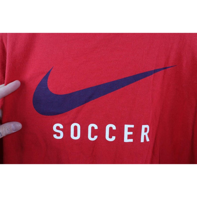 May include: Red t-shirt with a large navy blue Nike swoosh logo and the word "SOCCER" in white block letters. The shirt is a solid red color.