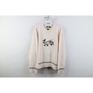 May include: A cream-colored long-sleeve sweater featuring a textured cable knit pattern throughout. The center of the chest displays an embroidered graphic of two birds perched on a branch with red berries. A dark leaf border accents the cuffs and hem.